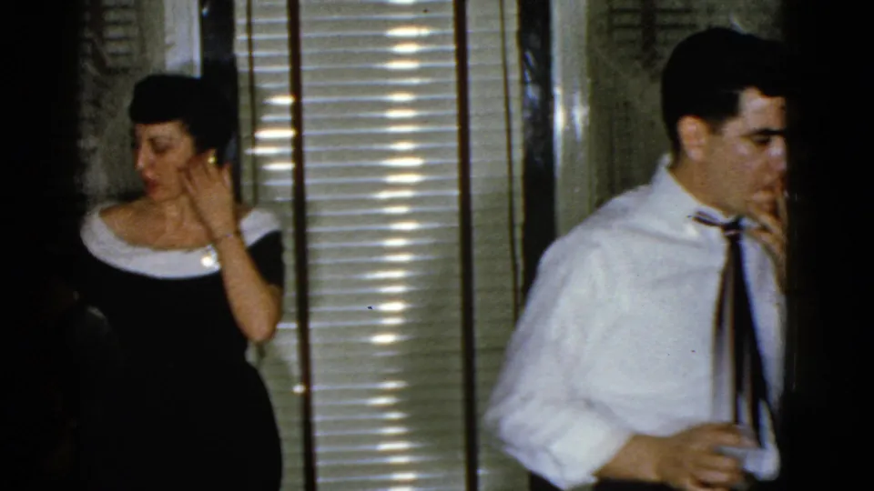 1975 Boston home-movie frame showing a woman in a dark dress near venetian blinds and a man in a white shirt holding a drink.