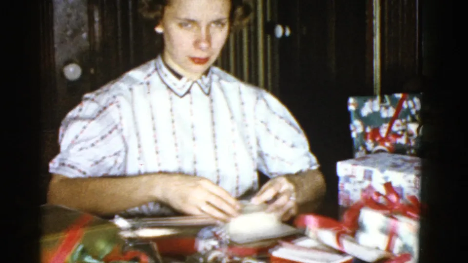In a 1975 Boston home-movie frame, a woman in a striped blouse folds paper at a table near wrapped gift boxes.