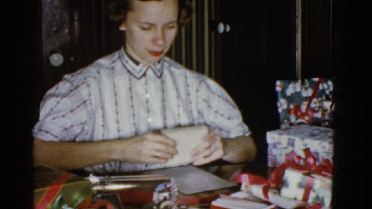 Adult woman in a striped blouse wraps a gift at a table in a Boston home-movie Christmas scene from 1975.