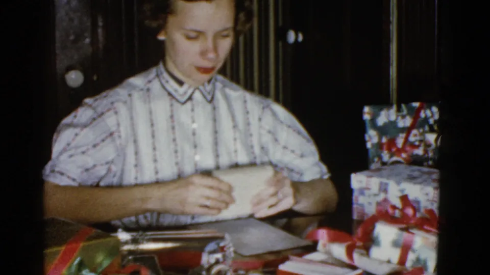 Adult woman in a striped blouse wraps a gift at a table in a Boston home-movie Christmas scene from 1975.