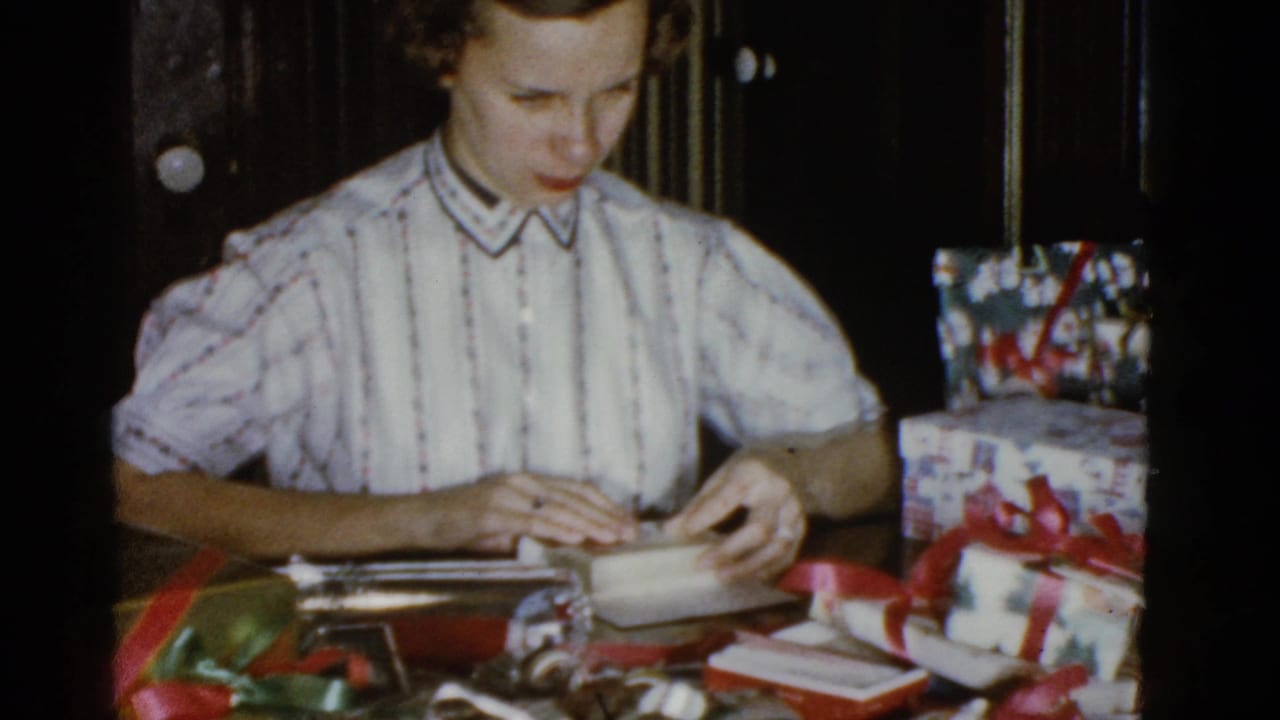 Adult woman concentrates on folding gift wrap at a table with presents and ribbon in a 1975 Boston home interior.