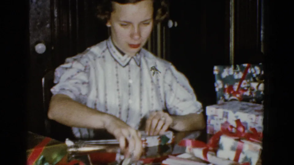 In a dim private interior, the woman reaches for supplies while wrapping Christmas presents on 8mm home-movie footage.
