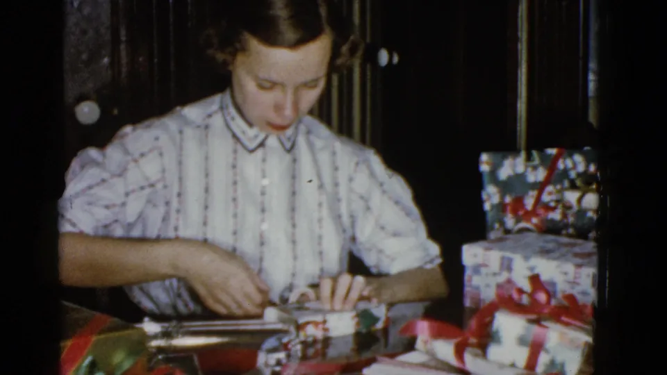 Adult woman presses wrapping material onto a small package, surrounded by ribbon and presents in a 1975 Boston holiday scene.