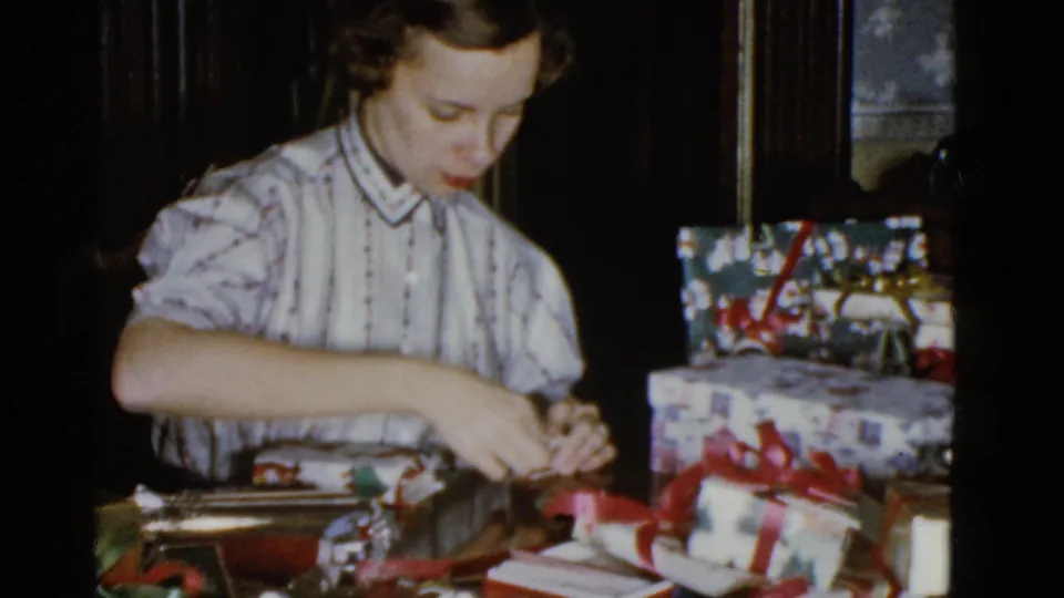 In a 1975 Boston home-movie frame, a woman in a striped blouse works on ribbon at a table with Christmas gifts nearby.