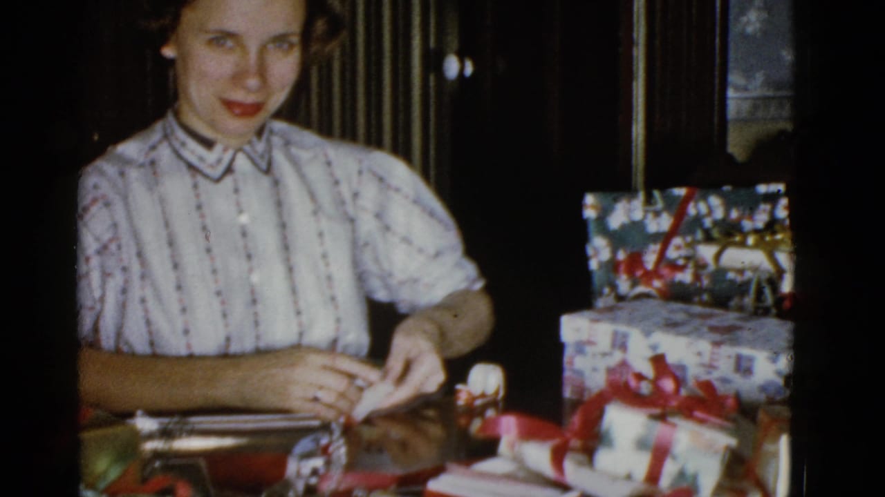 A woman smiles toward the camera while handling ribbon beside patterned Christmas packages in a private interior.