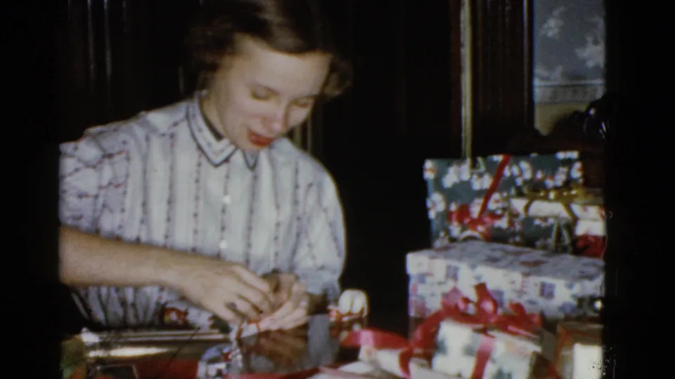 The woman looks down at her hands while tying ribbon, with stacked wrapped Christmas gifts visible beside her.