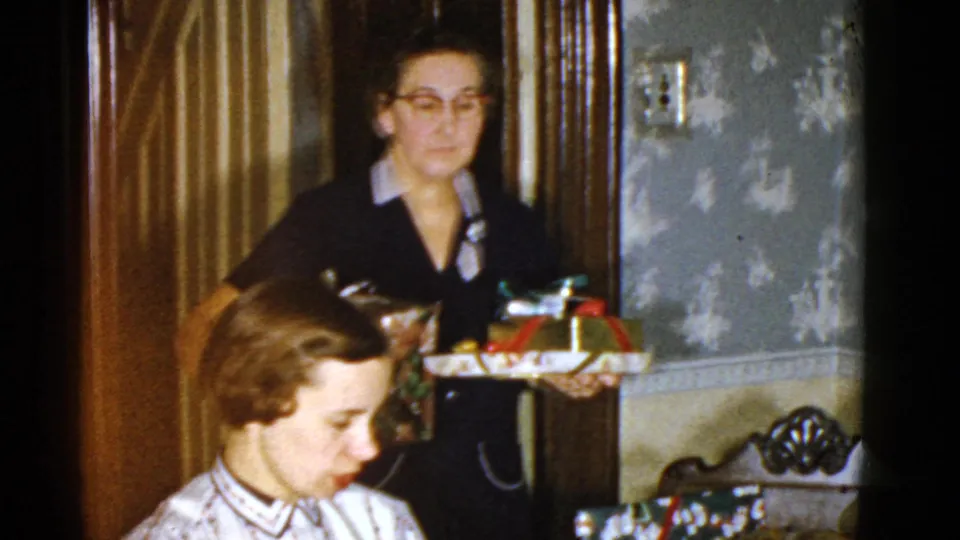 In a Boston home interior, a woman with glasses holds wrapped gifts behind a seated woman at the holiday table.