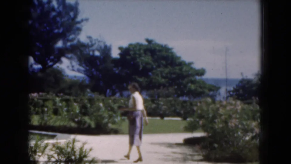 In 1975 Florida home-movie footage, a woman walks along a sunny garden path with hedges, trees, water, and skyline beyond.