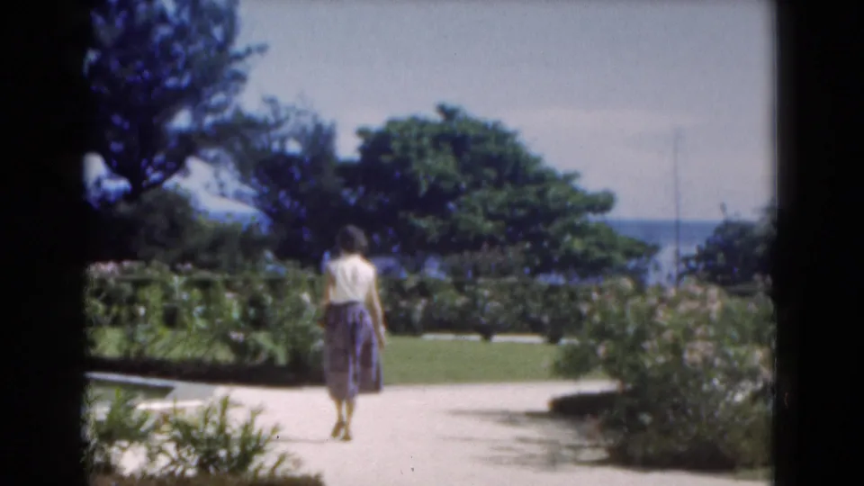A woman in a light top and patterned skirt walks away through a Florida garden, with trees, water, and a city skyline in the distance.