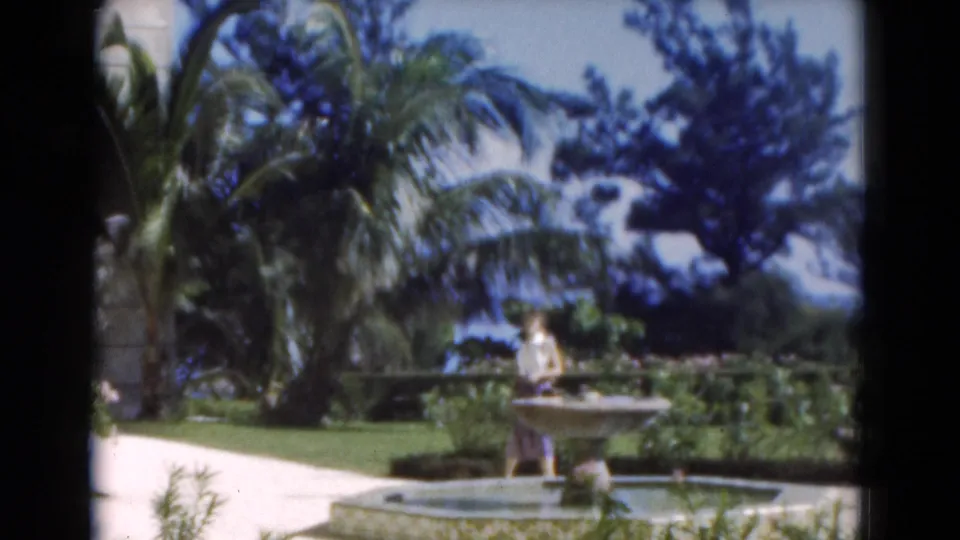 In 1975 Florida home-movie footage, a woman stands by a stone fountain in a sunlit garden with palm trees and manicured greenery.