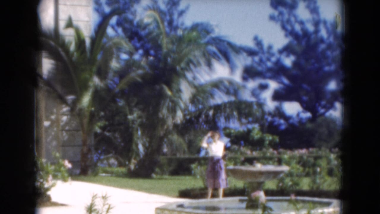 Woman waving beside a fountain in a palm-filled garden.