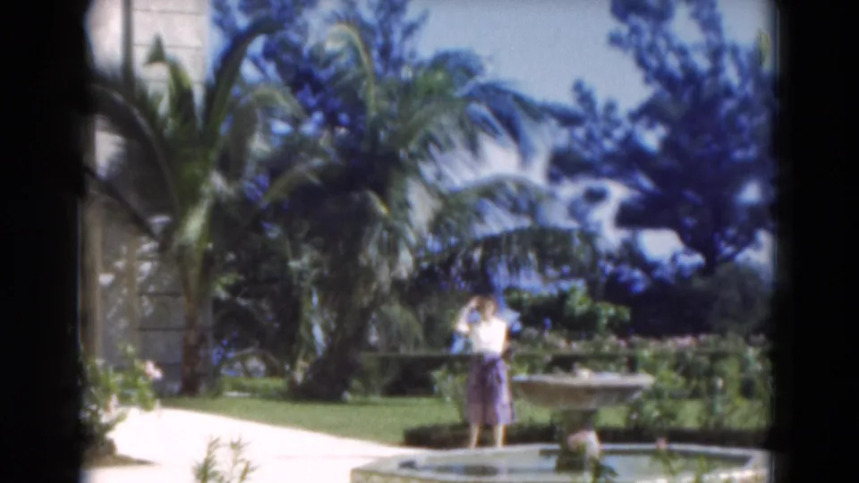 A woman in a light top and purple skirt waves toward the camera beside a stone fountain in a Florida garden, 1975.