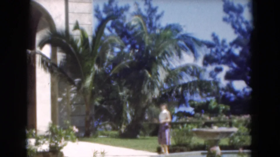 The woman walks through the 1975 Florida garden scene, passing palms, shrubs, and a stone fountain in bright daylight.