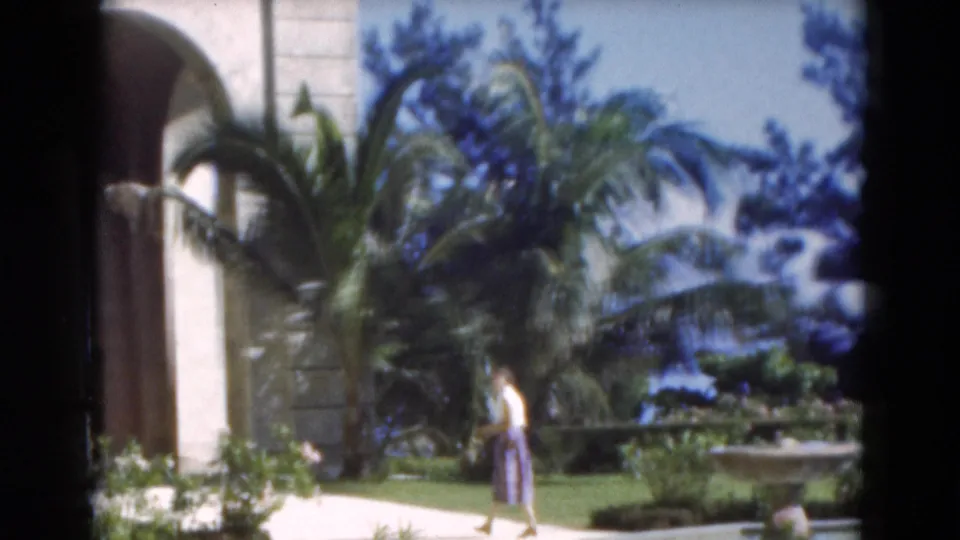 The woman moves past a building edge and palms near the fountain in sunlit 1975 Florida 8mm home-movie footage.