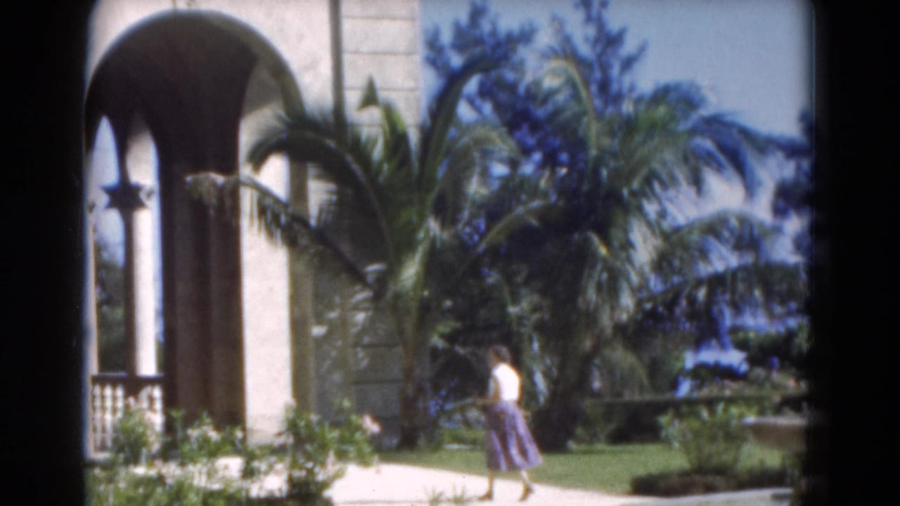 Woman in a white top walks near an arched building and palm trees.