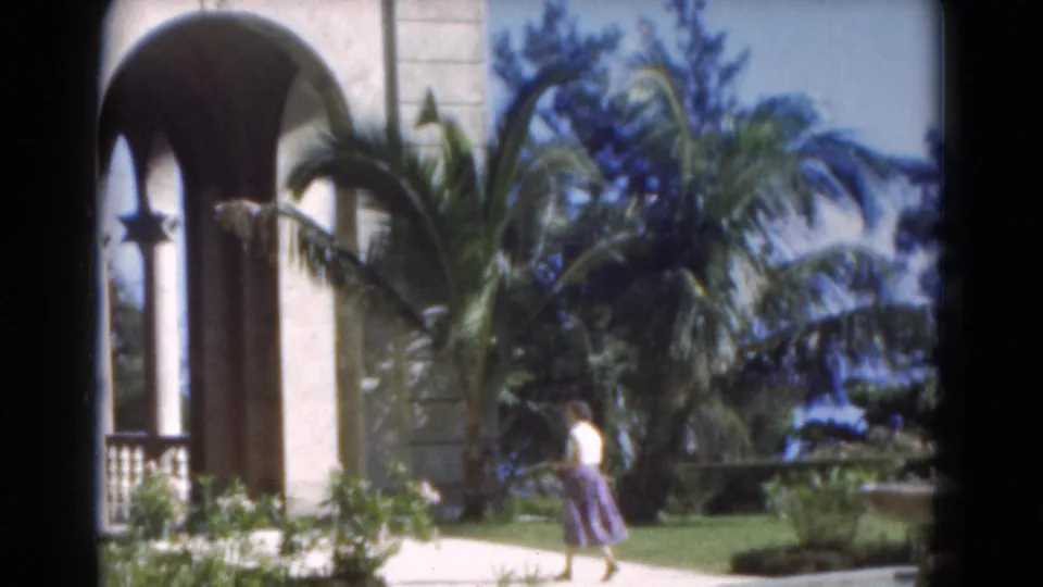 1975 Florida 8mm frame of a woman walking along a bright garden path beside palms and arched stucco architecture.