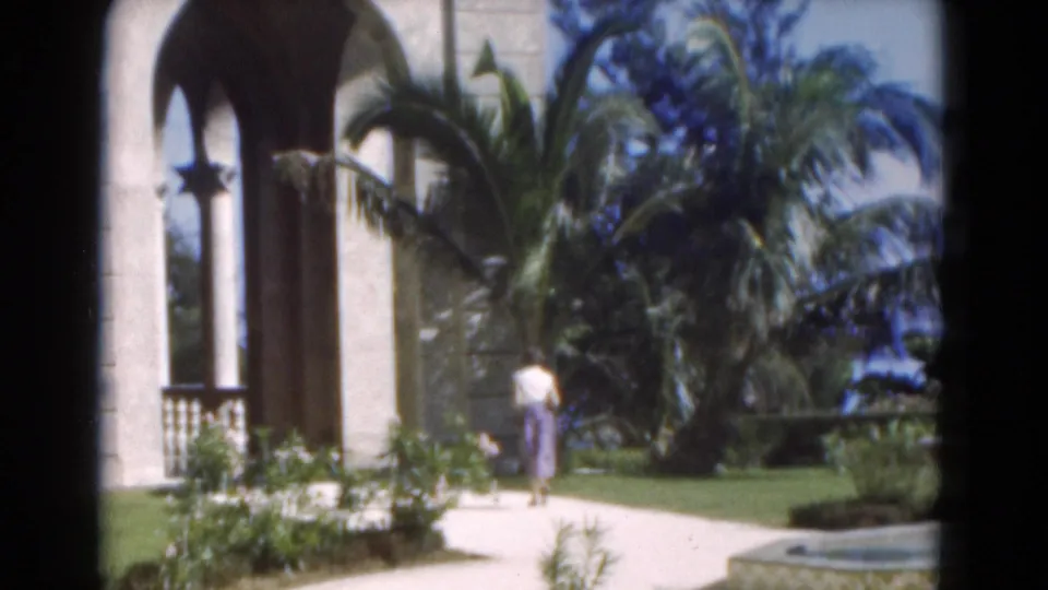 A 1975 home-movie frame shows the woman continuing through the tropical garden beside an archway, palms, lawn, and low plantings.