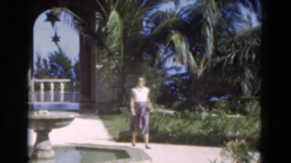 The woman appears near a stone fountain in the Florida garden, with palms, an arched entry, lawn, and sunlit path visible.