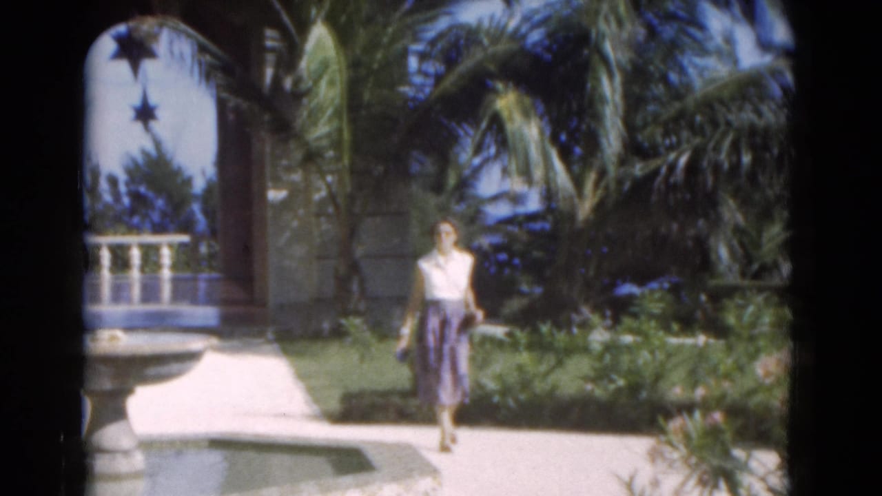 In 1975 Florida 8mm footage, a woman in a white blouse and blue skirt walks through a tropical garden near a stone fountain and palm trees.
