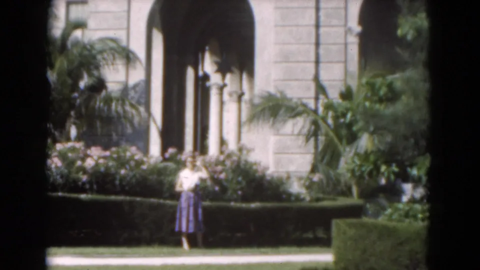 In front of an arched-column building in Florida, the woman stands near a low hedge with palm fronds and flowering shrubs around her.