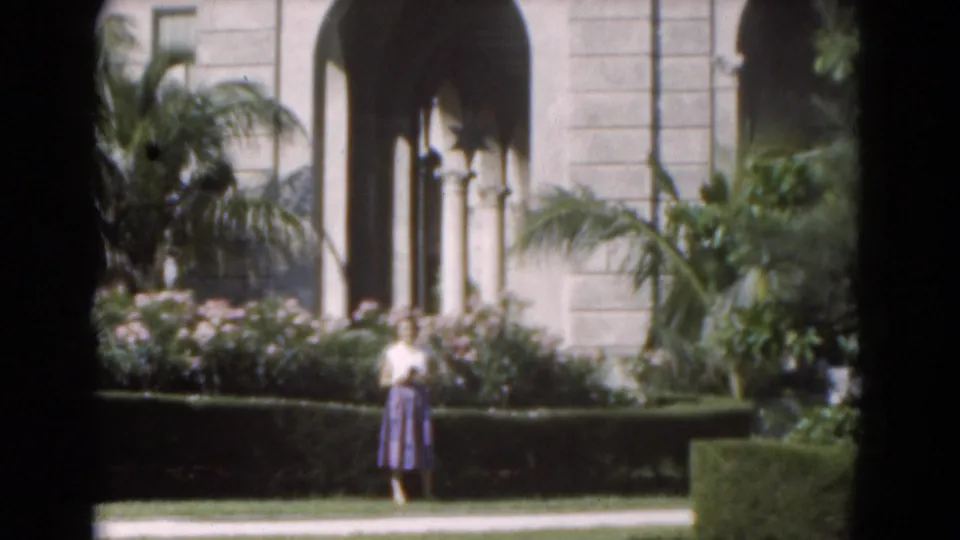 The woman remains near a low hedge in front of a sunlit arched entrance, with tropical planting visible across the garden foreground.
