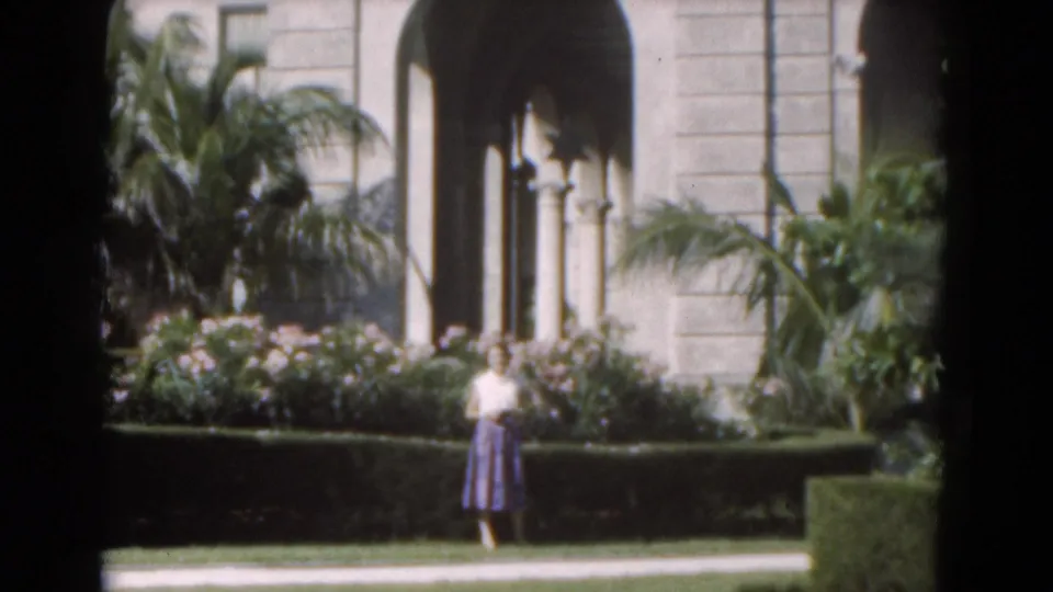 1975 Florida 8mm frame showing a woman near a manicured garden, palms, flowering shrubs, and arched building columns.