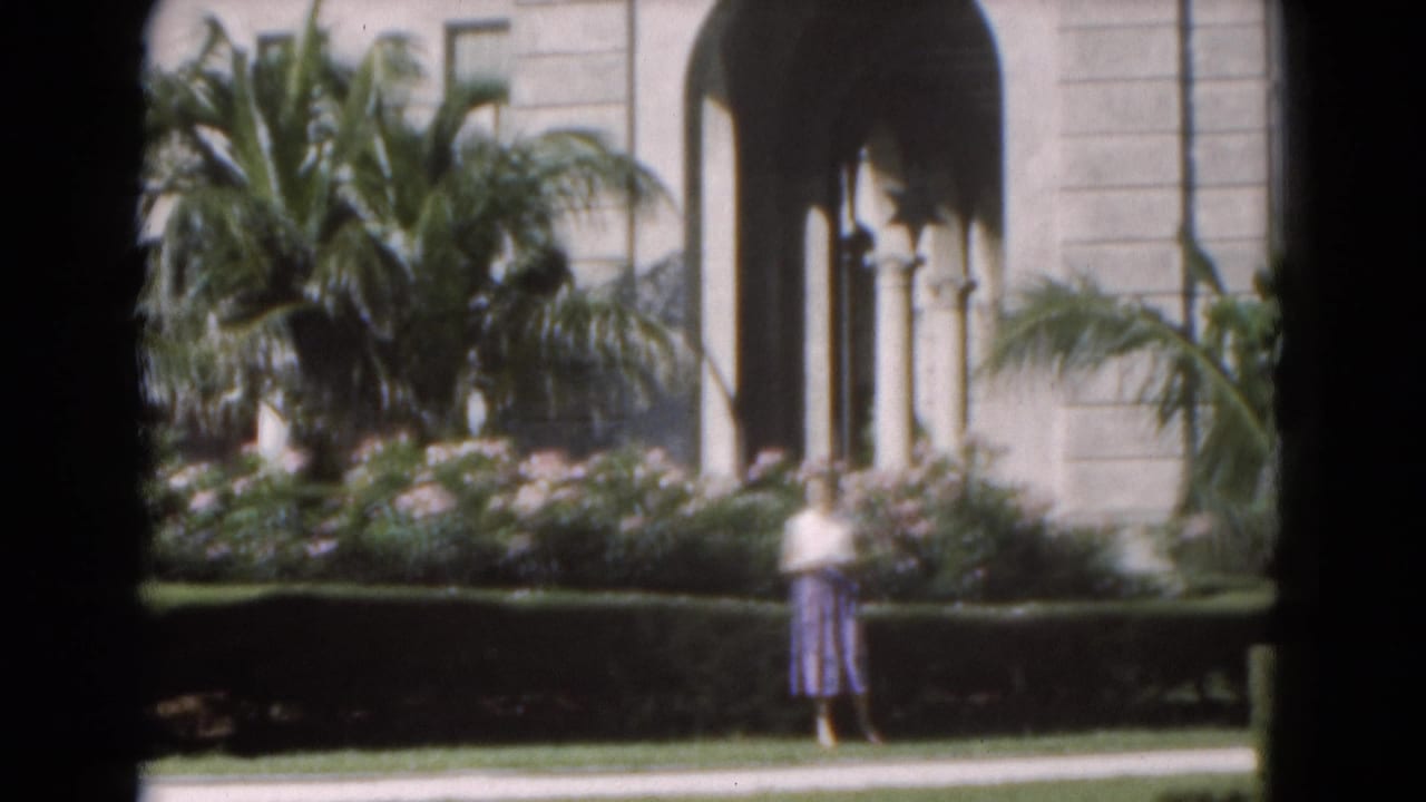 Woman walking beside a hedge with palms and arched colonnades behind her.