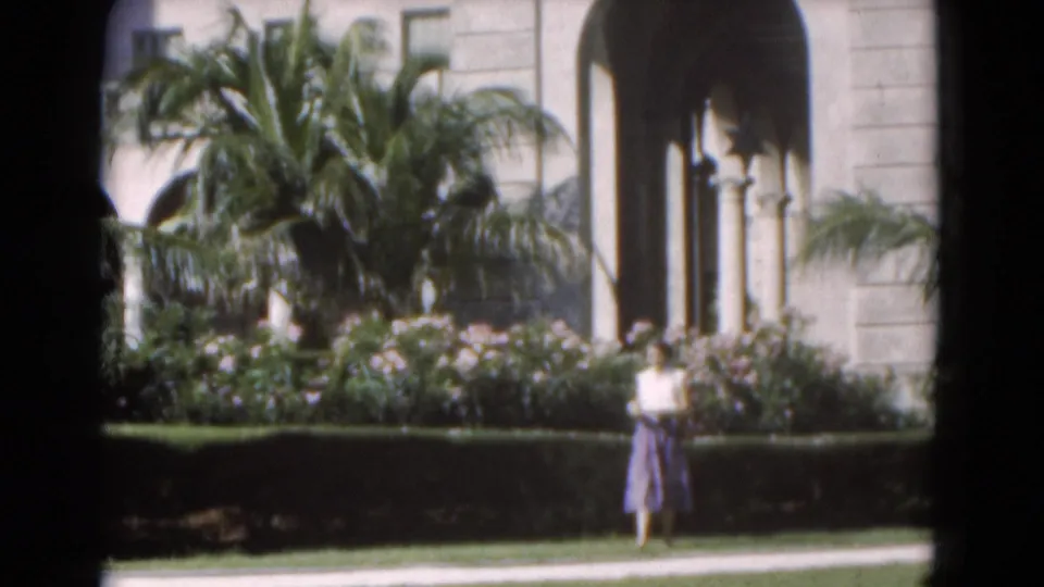 Soft 8mm frame from Florida in 1975 showing a woman in a garden foreground below palms and an arched building exterior.