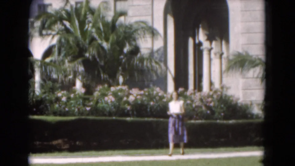 1975 Florida 8mm still with a woman centered in a landscaped garden before palms, flowers, hedges, and arched architecture.
