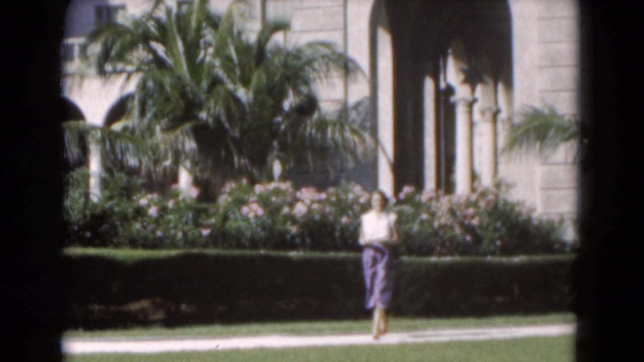 Woman walking on a path beside palms, flowers, and arched stone columns.