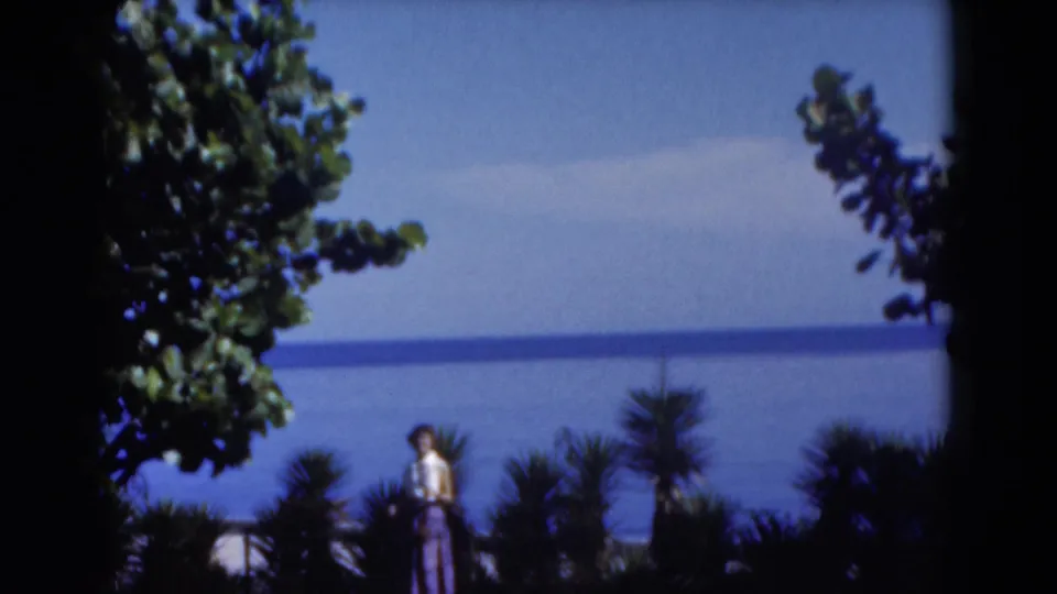1975 Florida 8mm frame of a woman in a tropical garden area, partly framed by trees, with calm blue ocean beyond.