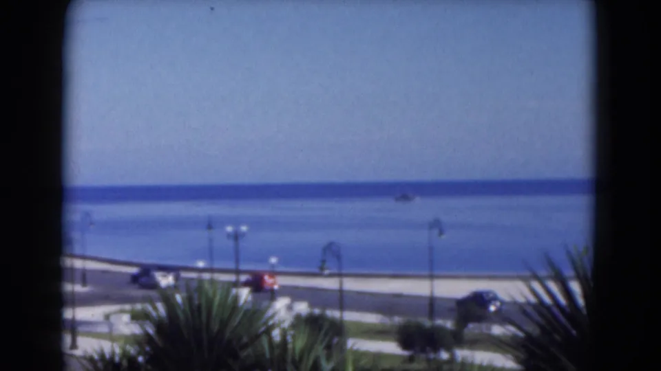 1975 Florida 8mm frame looking over palm foliage toward a coastal road, vintage cars, lampposts, and the ocean horizon.