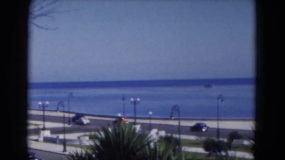1975 8mm Florida travel view over a coastal boulevard with cars, palms, lamps, and calm blue water beyond.