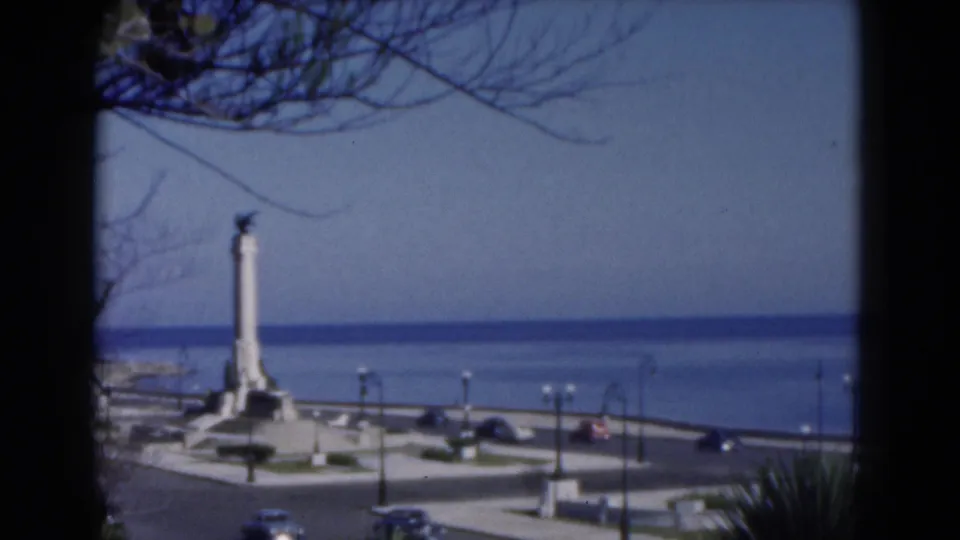 1975 Florida 8mm view of a white waterfront monument near moving cars, street lamps, palm plants, and open water.