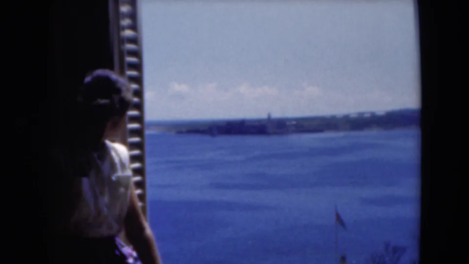 1975 Florida 8mm frame of a woman at a window with open blue water, shoreline, and hazy sky beyond.