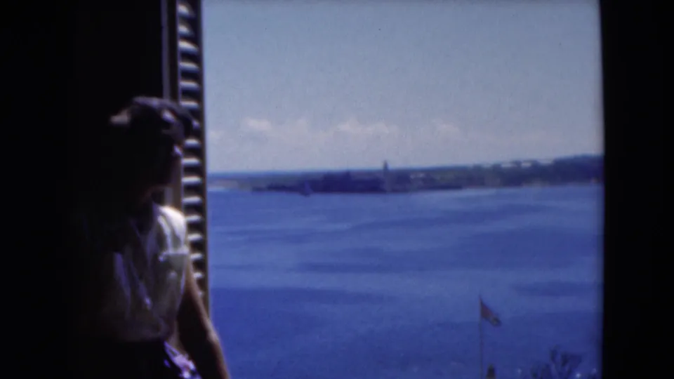 1975 Florida home-movie frame showing a woman silhouetted by a window while looking out over sea and low coastal land.
