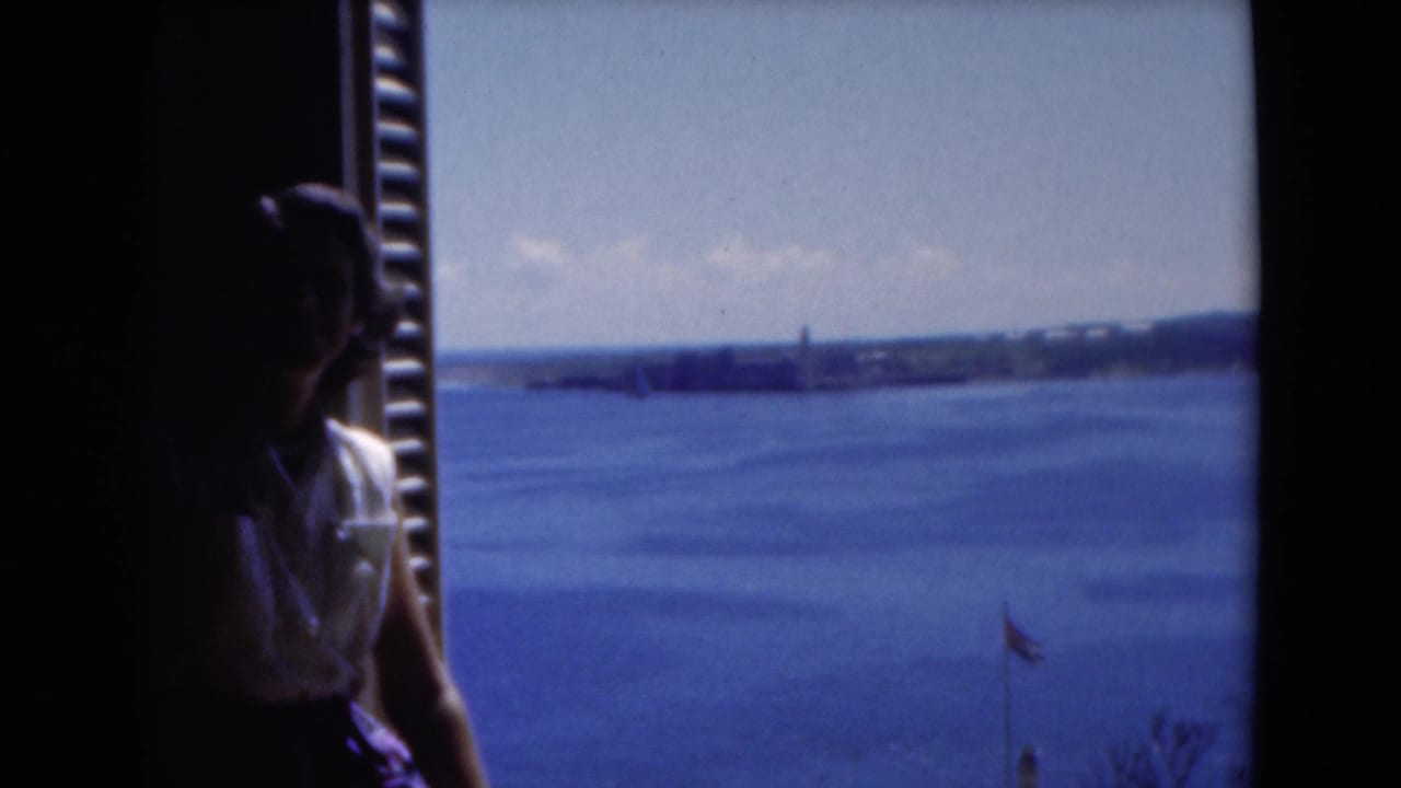 In 1975 Florida home-movie footage, a woman sits in shade by a window overlooking blue water and a distant shore.