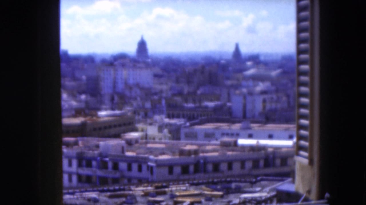 Elevated city view framed by shutters, with rooftops and domes beneath a cloudy sky.