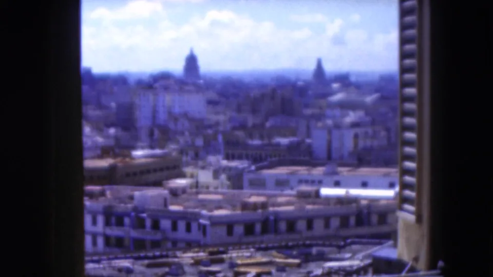 1975 Florida home-movie frame with an elevated, shutter-framed city view, low buildings, distant domes, and a bright partly cloudy sky.