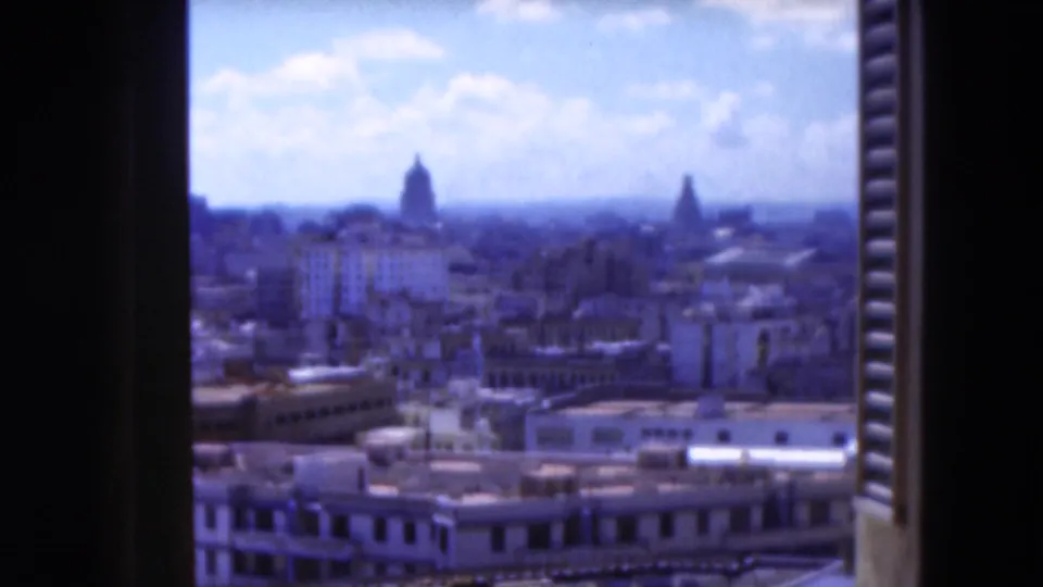 1975 Florida 8mm frame showing a soft, blue-toned city panorama through shutters, with low rooftops and domed forms on the skyline.