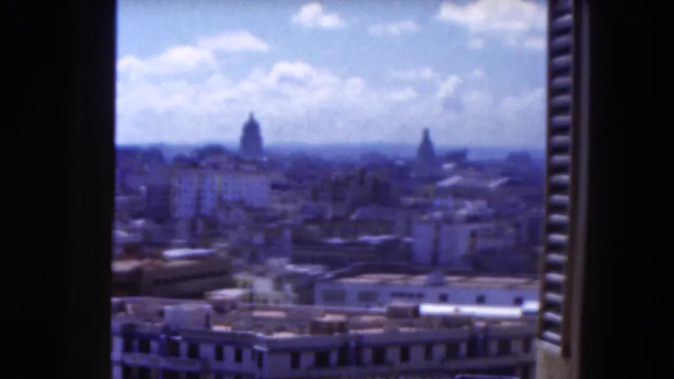 1975 Florida home-movie frame looking from a dark window opening over a hazy city skyline with domes, rooftops, and bright clouds.