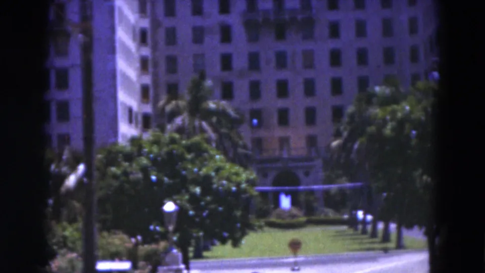 1975 Florida 8mm frame showing a towered building facade beyond a landscaped lawn, trees, palm fronds, and small lamps.