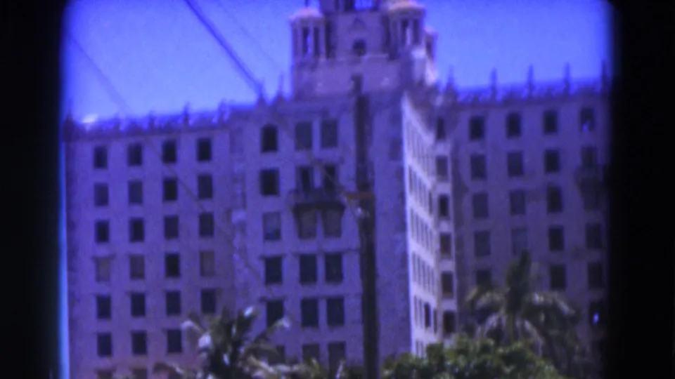 1975 Florida 8mm frame showing a white building facade, many windows, palms, blue sky, and a rooftop tower feature.