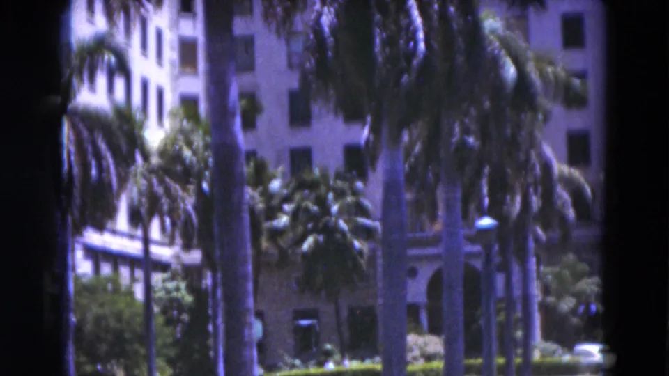 1975 Florida 8mm frame of palms, shrubs, a lamp post, and the lower facade of a white resort-like building.