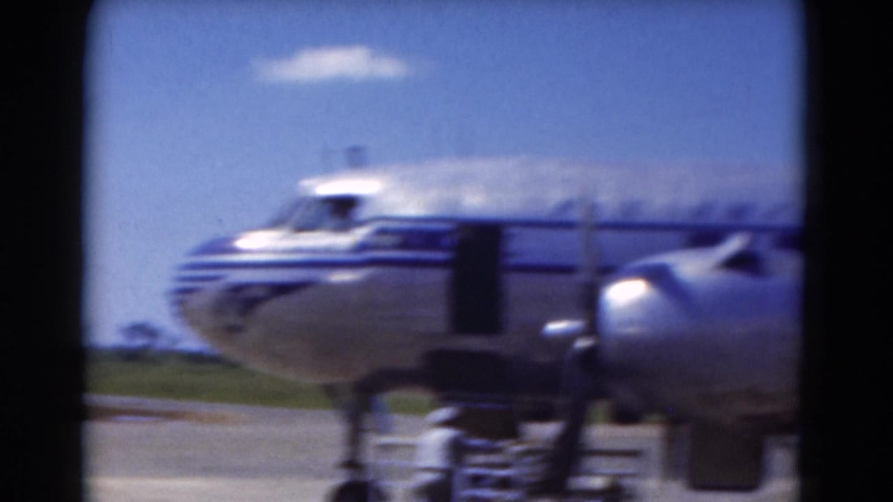 Silver propeller airplane with blue stripes parked on a sunny airport tarmac.