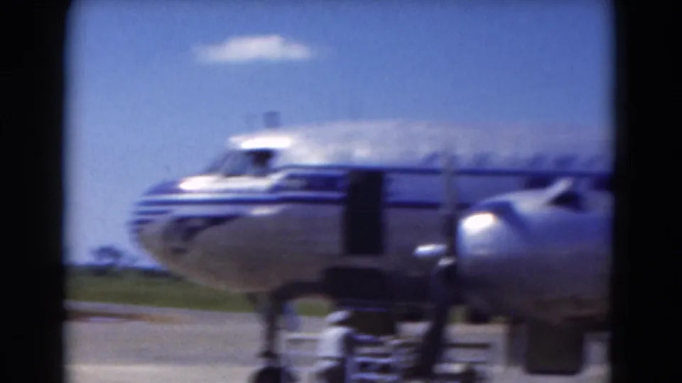 1975 Florida 8mm frame showing the nose and propeller area of a silver airplane with blue stripes on the tarmac.
