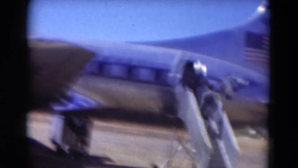 Soft 1975 Florida home-movie frame showing the aircraft side, mobile stairs, and a partial American flag on the tail.