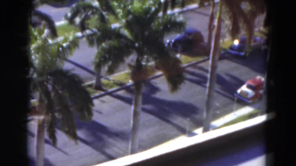 Elevated 1975 Florida home-movie view with a red car at the curb, palm trees, streetlights, pavement, lawn, and a bright balcony edge.