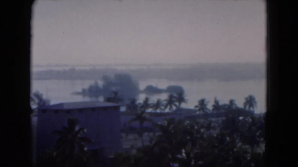 1975 Florida 8mm frame showing palms, a low building, water, and distant shoreline through a window.