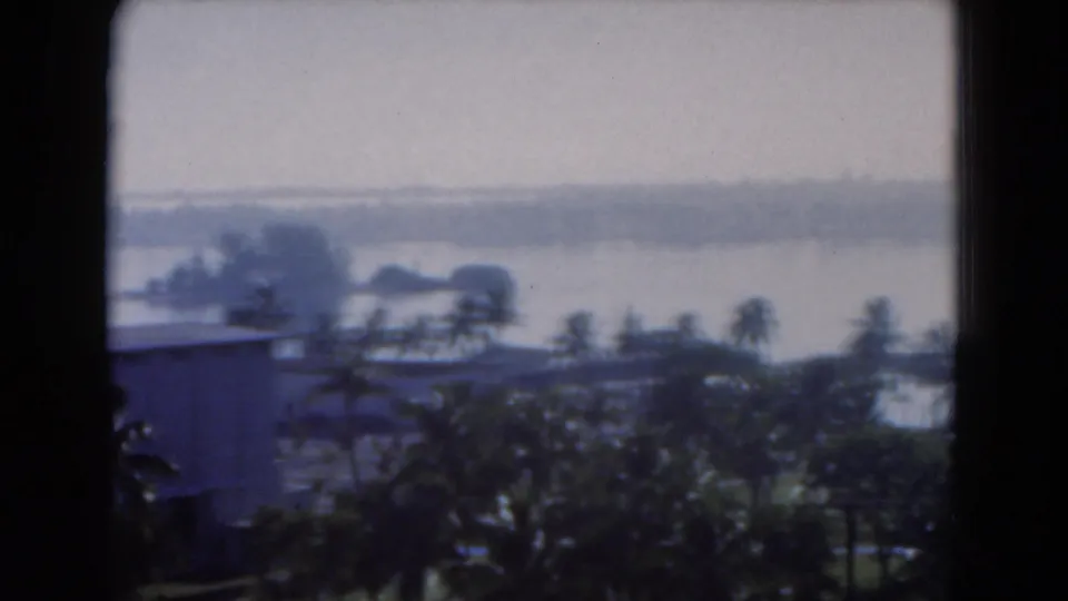 1975 Florida scene with a low building, tropical trees, lawns, and a distant shoreline viewed through glass or a window.